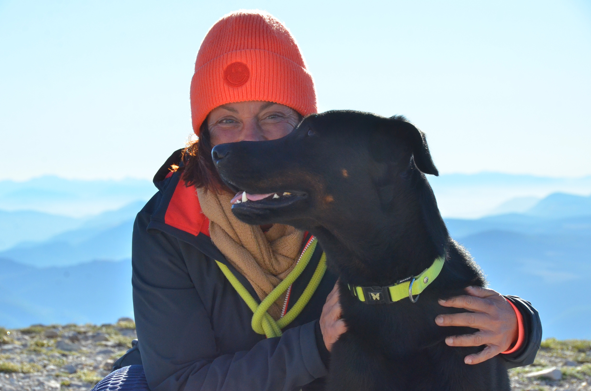 Une personne au bonnet rouge enlace un grand chien noir.
